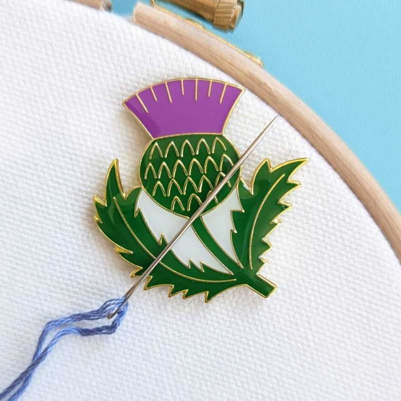An embroidery needle rests on a Scottish thistle magnet, on a white fabric background.