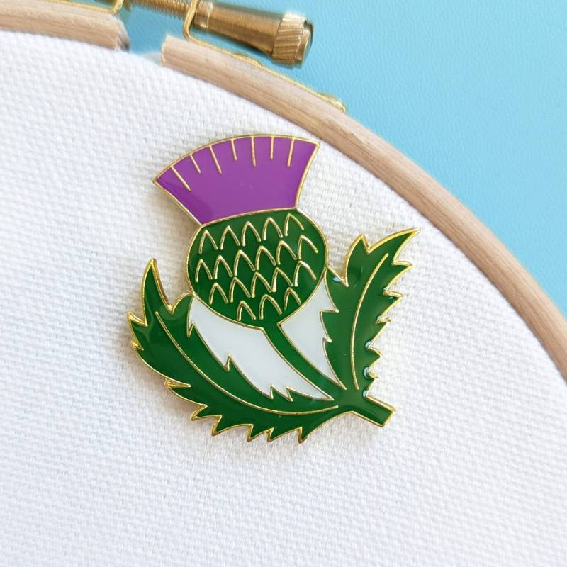An embroidery needle rests on a Scottish thistle magnet, on a white fabric background.