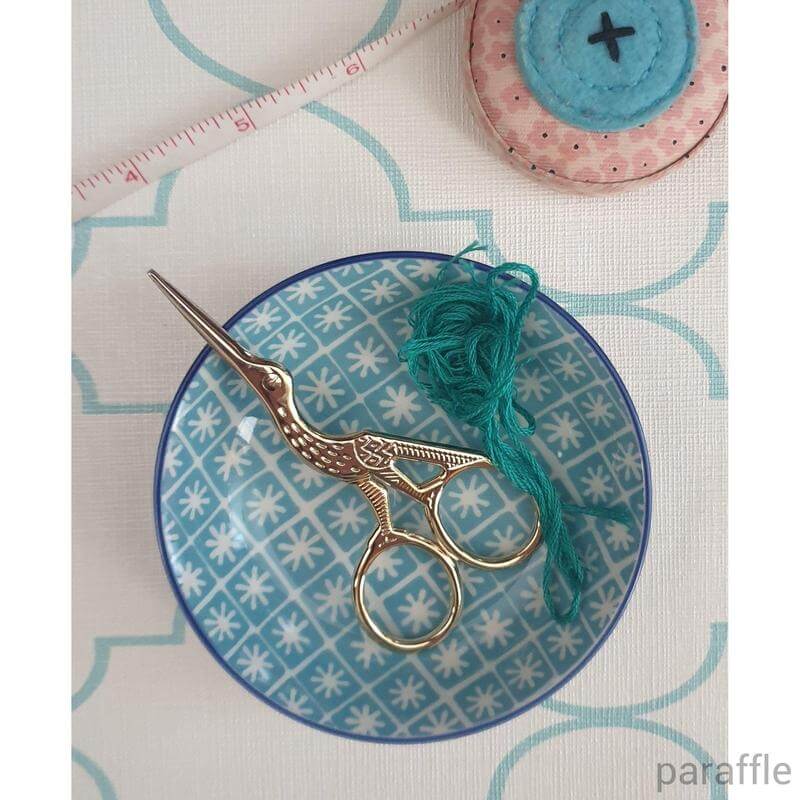 A pair of stork embroidery scissors resting in a small patterned bowl containing teal embroidery thread.
