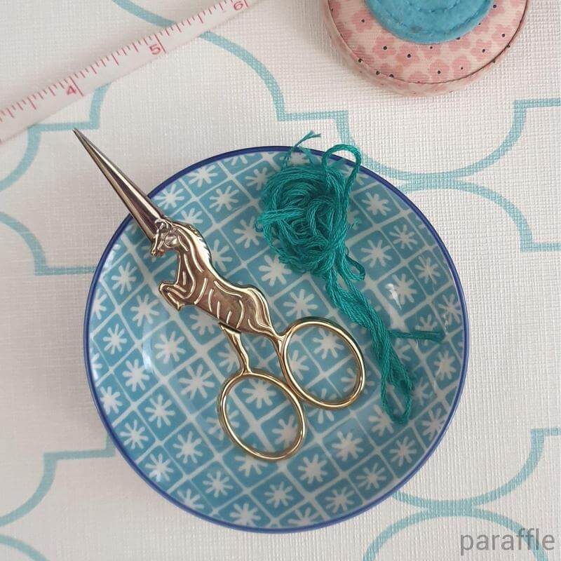A pair of gold unicorn embroidery scissors resting in a bowl with some turquoise embroidery thread.
