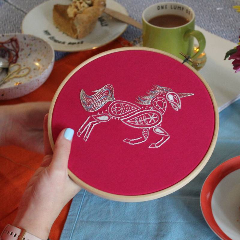 A pair of hands holding an embroidery hoop containing a piece of bright pink fabric. The fabric is embroidered with a white unicorn design.